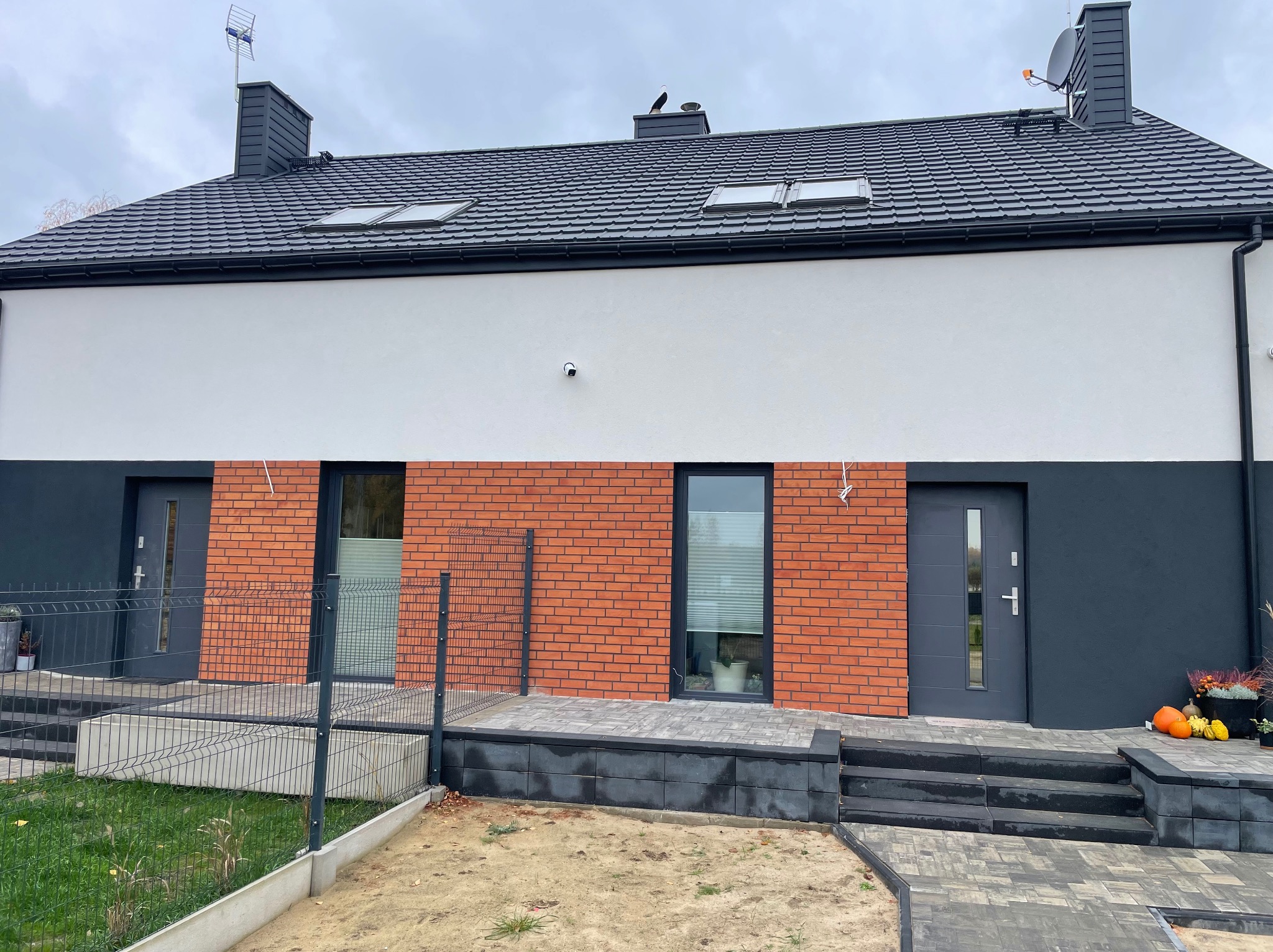 Nowoczesny dom jednorodzinny z elewacją w kolorach bieli, szarości i czerwonej cegły. Dach z oknami połaciowymi, antena satelitarna i komin.