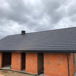 Dom w trakcie budowy z ciemną dachówką ceramiczną ułożoną na gotowej konstrukcji dachu, widoczny komin i rynny, ściany z czerwonego ceglanego pustaka.