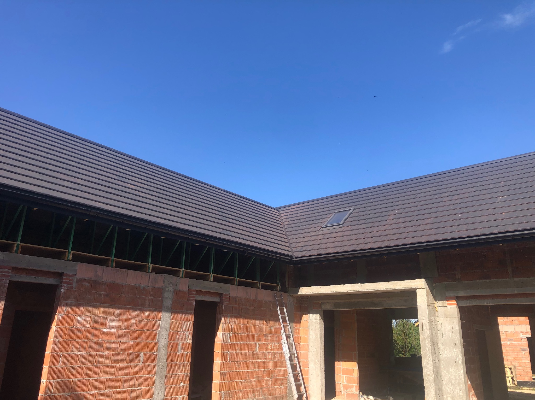 Częściowo ukończony budynek z cegły z ciemnobrązowym dachem i oknem dachowym w słoneczny dzień, drabina oparta o ścianę.