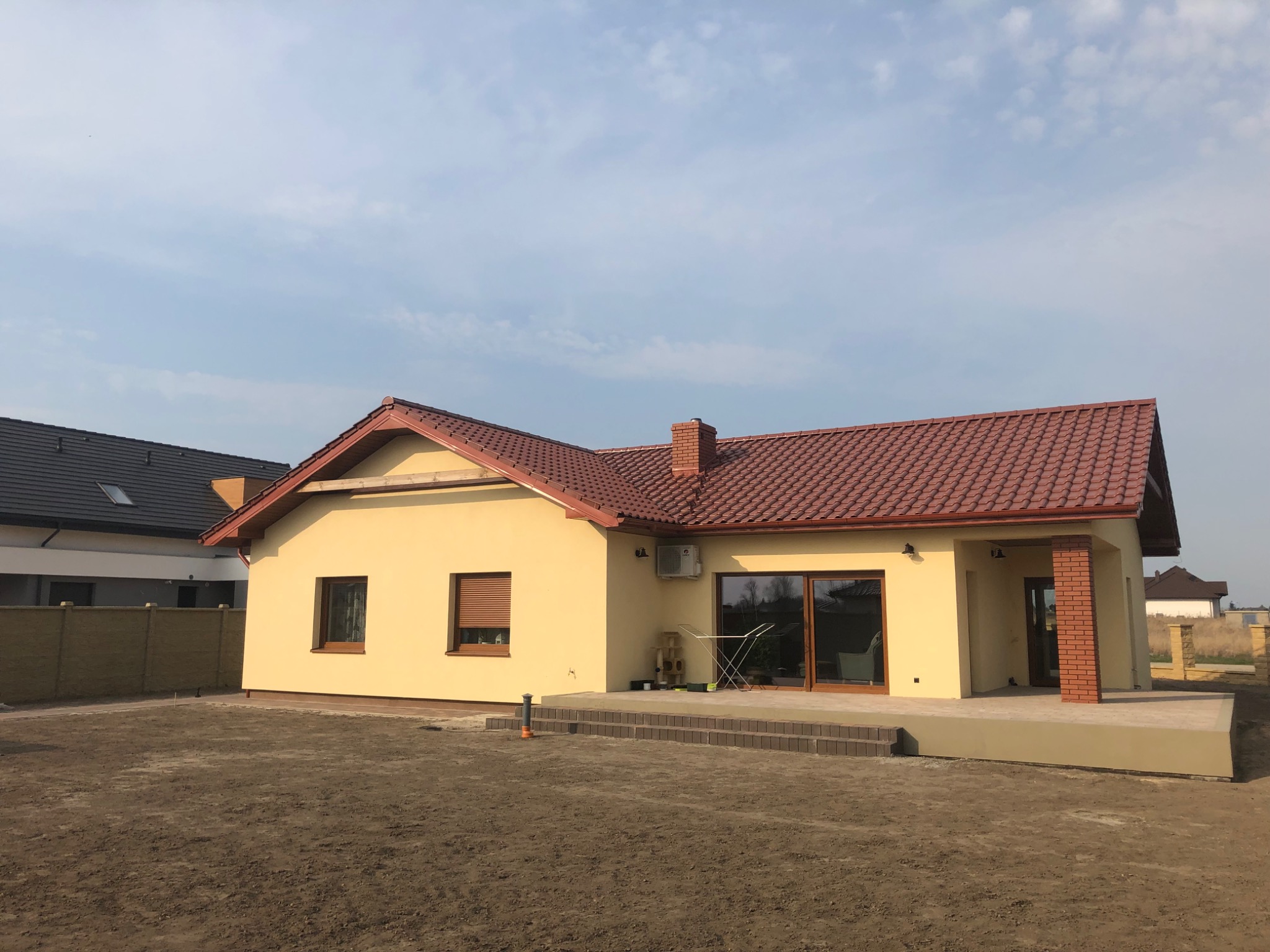 Żółty dom jednorodzinny z czerwoną dachówką, ceglanym kominem i kolumną, widok z przodu na niezagospodarowaną działkę.