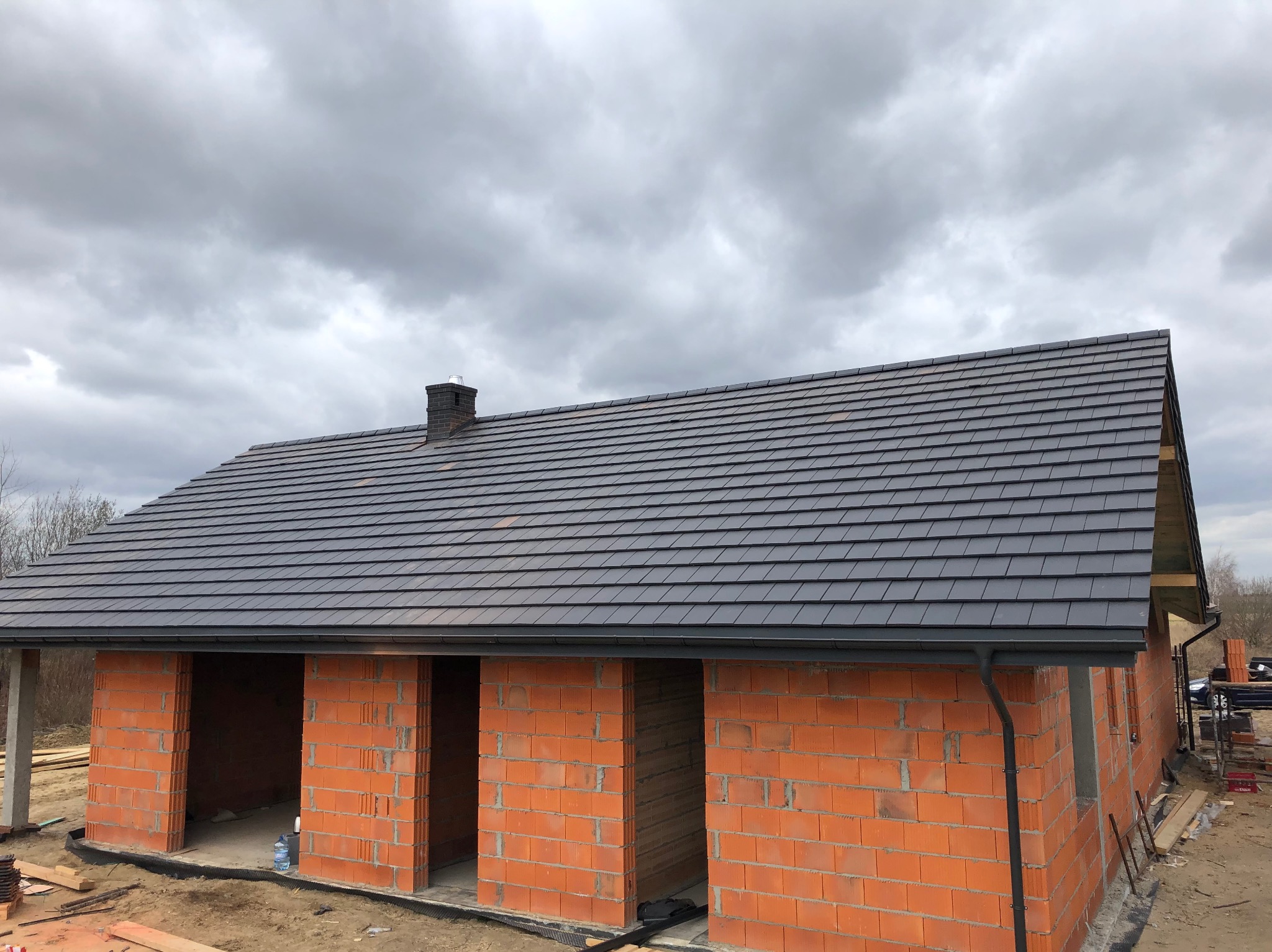 Dom w trakcie budowy z ciemną dachówką ceramiczną ułożoną na gotowej konstrukcji dachu, widoczny komin i rynny, ściany z czerwonego ceglanego pustaka.