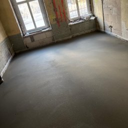 DoMix - Świeżo wylana, gładka posadzka jastrychowa w surowym wnętrzu z dwoma oknami i odsłoniętymi cegłami, widoczne ślady po remoncie na ścianach.