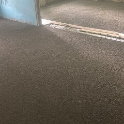 DoMix - Świeżo wylana, ciemnoszara posadzka w pomieszczeniu w trakcie remontu. Widoczne niedoskonałości ścian i futryny drzwi. Listwy poziomujące na powierzchni posadzki.