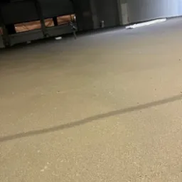 Wnętrze hali przemysłowej z nową, gładką posadzką w odcieniach beżu, widoczne ślady po szlifowaniu, w tle brama wjazdowa i fragment ściany.