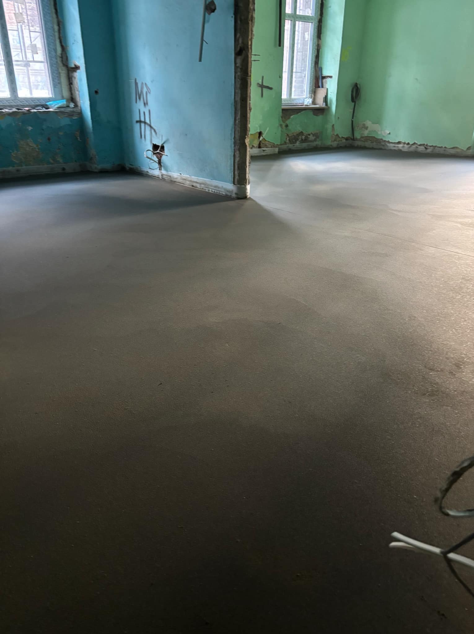 Świeżo wylana, gładka posadzka w pomieszczeniu z fragmentami ścian w kolorach niebieskim i zielonym, widoczne ślady po instalacjach i niedokończone wykończenie ścian.