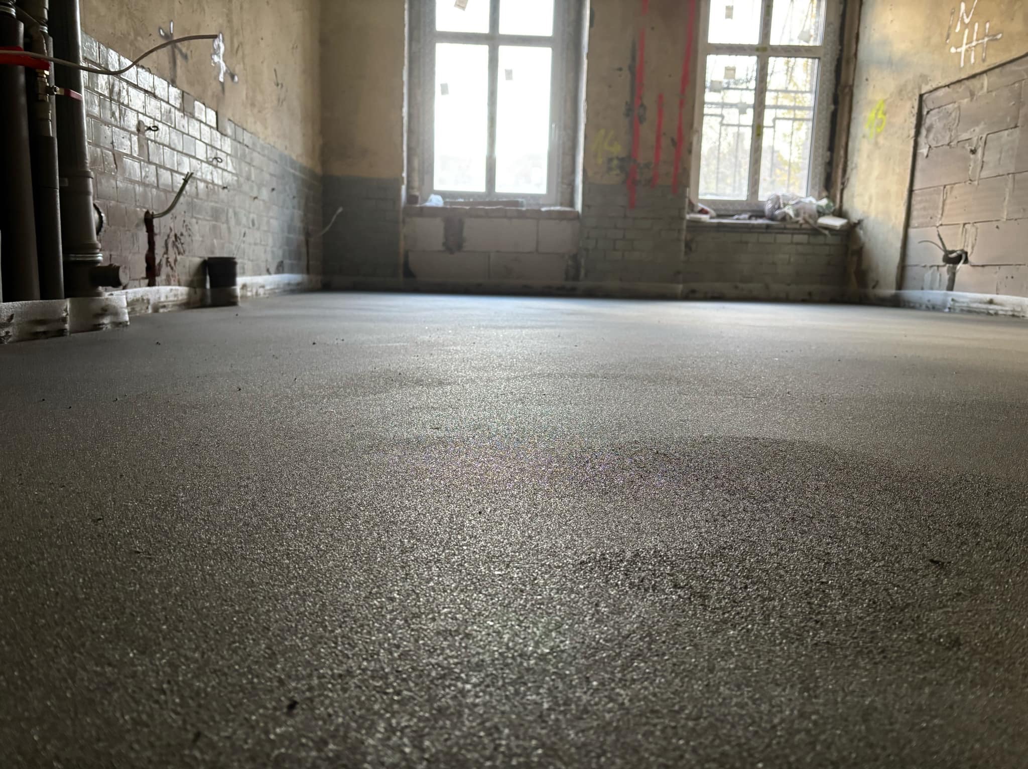 Świeżo wylana, gładka, szara posadzka w surowym wnętrzu z widocznymi instalacjami i dwoma oknami z parapetami wyłożonymi cegłami.