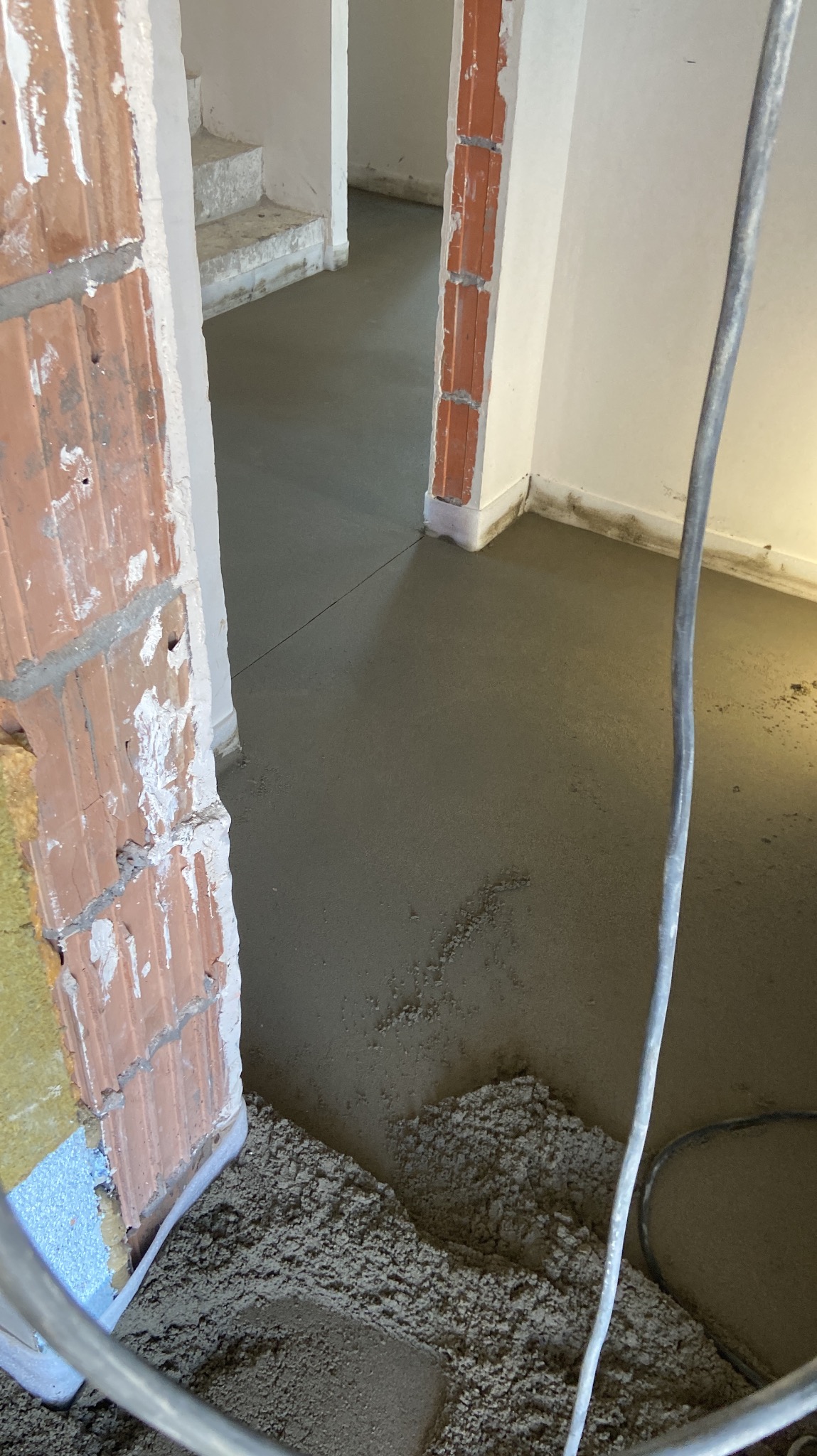 Świeżo wylana, gładka, szara posadzka w surowym wnętrzu z widocznymi elementami muru z cegły i kablem elektrycznym w kadrze.