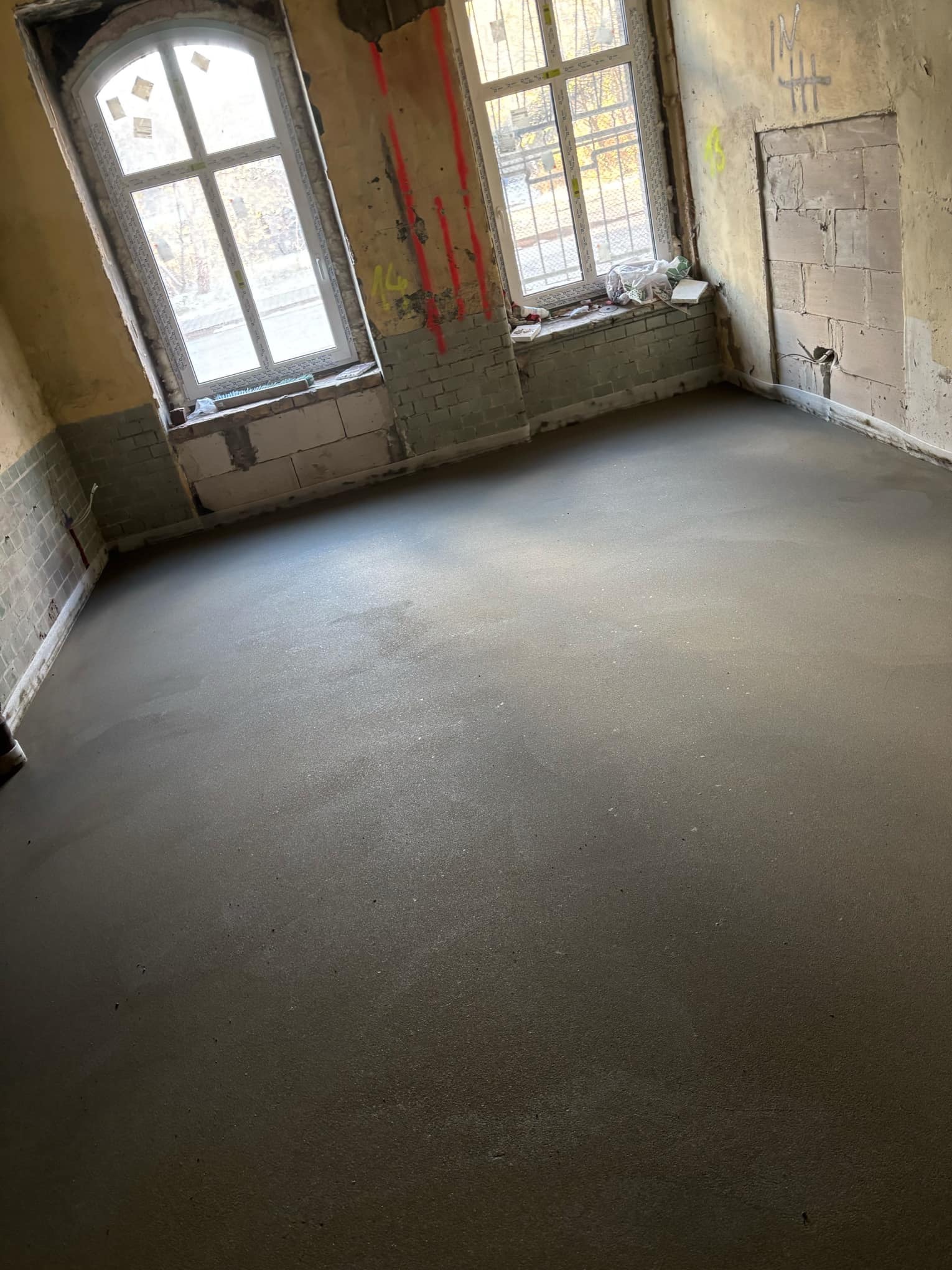Świeżo wylana, gładka posadzka jastrychowa w surowym wnętrzu z dwoma oknami i odsłoniętymi cegłami, widoczne ślady po remoncie na ścianach.