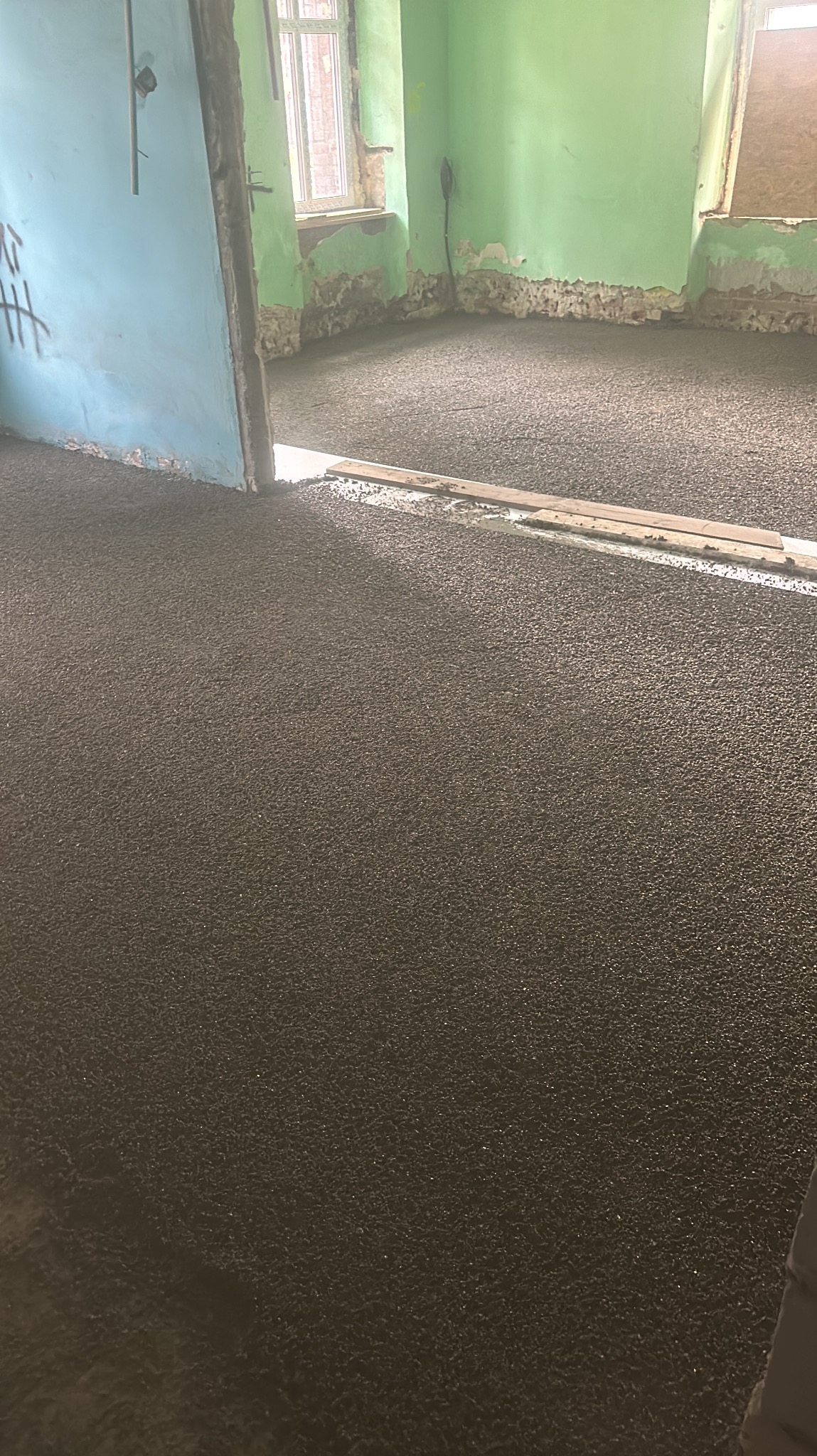 Świeżo wylana, ciemnoszara posadzka w pomieszczeniu w trakcie remontu. Widoczne niedoskonałości ścian i futryny drzwi. Listwy poziomujące na powierzchni posadzki.