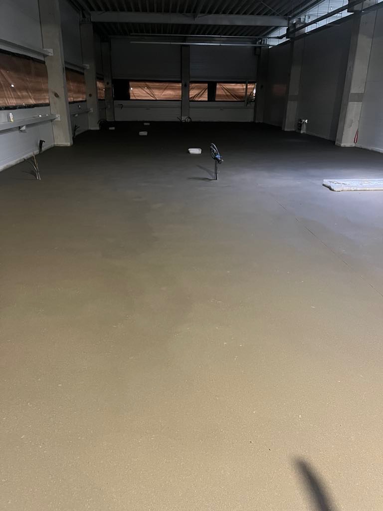 Świeżo wylana, gładka posadzka w hali przemysłowej z widocznymi elementami instalacji wystającymi z podłoża i oknami w tle.