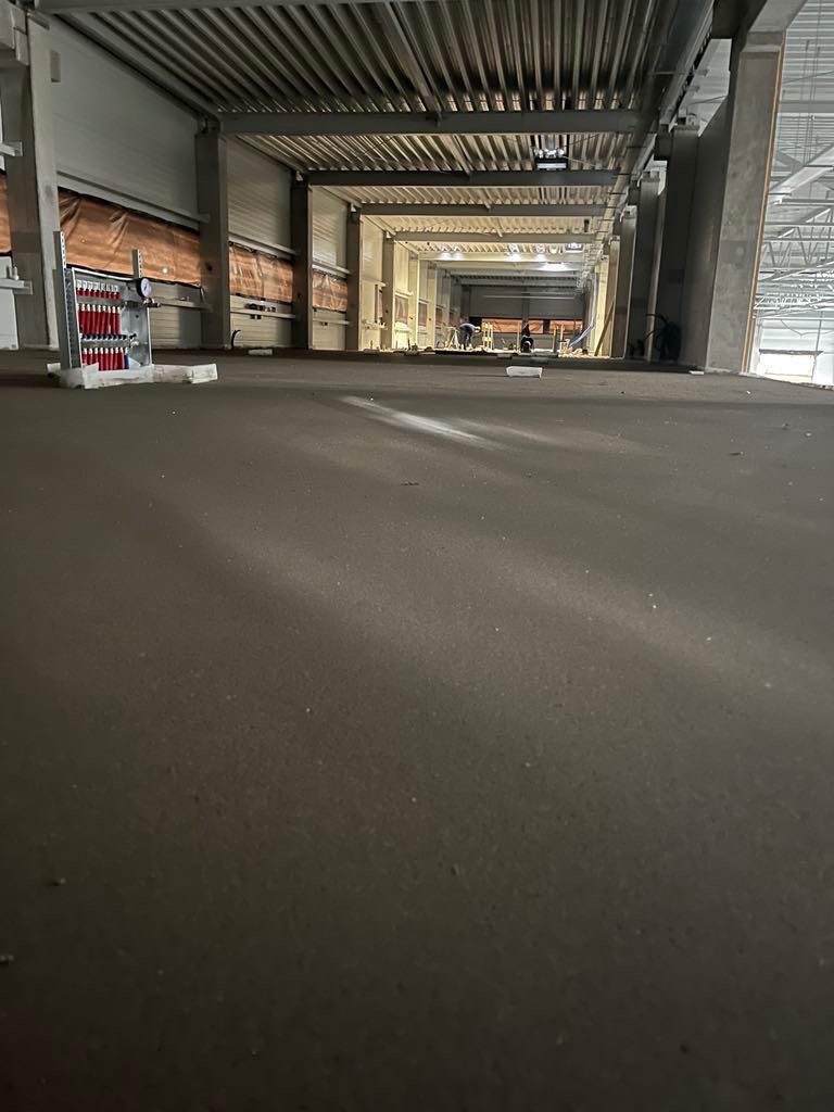 Świeżo wylana, ciemnoszara posadzka w hali przemysłowej, widok z niskiego kąta, w tle widoczne osoby pracujące i instalacje.
