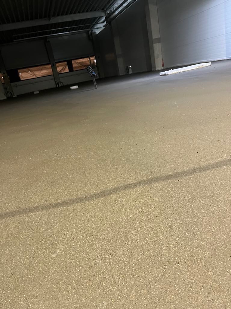 Wnętrze hali przemysłowej z nową, gładką posadzką w odcieniach beżu, widoczne ślady po szlifowaniu, w tle brama wjazdowa i fragment ściany.