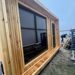 Monsal - Drewniana konstrukcja z dużym oknem i małym wejściem, prawdopodobnie w trakcie budowy, widoczny pracownik malujący elementy obok, na zewnątrz, pochmurna pogoda.