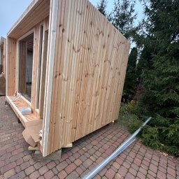 Monsal - Surowa, drewniana konstrukcja w trakcie budowy, z widocznymi elementami łączeniowymi i listwami, umieszczona na kostce brukowej przed zielonymi drzewami iglastymi.