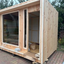 Monsal - Surowa drewniana konstrukcja z dużym oknem i otwartymi drzwiami, widoczne materiały izolacyjne wewnątrz, kabel i skrzynka elektryczna na zewnątrz, na kostce brukowej.