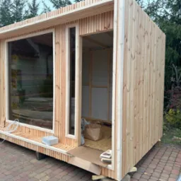 Surowa drewniana konstrukcja z dużym oknem i otwartymi drzwiami, widoczne materiały izolacyjne wewnątrz, kabel i skrzynka elektryczna na zewnątrz, na kostce brukowej.