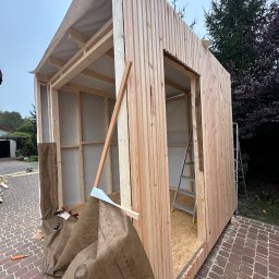 Monsal - Budowa drewnianej konstrukcji w Łazach, prawdopodobnie w trakcie wznoszenia, z widocznymi elementami szkieletu i częściowym obłożeniem deskami. Na pierwszym planie narzędzia i materiały budowlane.