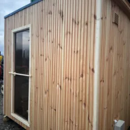 Nowoczesna, drewniana konstrukcja z pionowymi listwami, prawdopodobnie przenośna, z widocznym oknem i elementami wentylacji, ustawiona na zewnątrz na tymczasowych podporach.