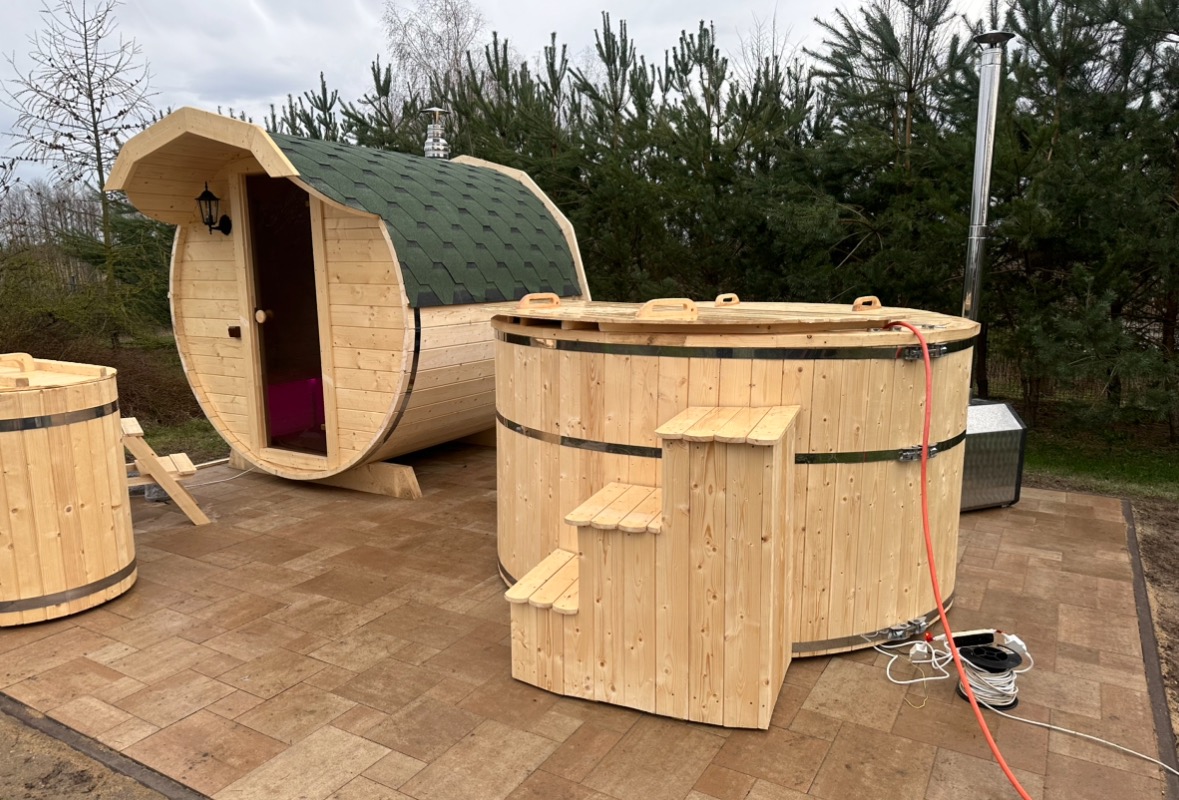 Drewniana beczka z zielonym dachem i lampą obok drewnianej balii z schodkami na zewnątrz, otoczone drzewami iglastymi.