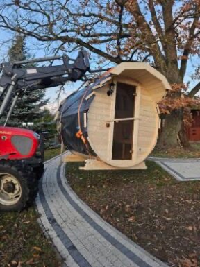 Transport beczki z jasnego drewna z drzwiami i lampą, prawdopodobnie do użytku jako miejsce relaksu, za pomocą traktora z czarnym mocowaniem, widok na zielony trawnik, drzewa i szarą kostkę brukową.
