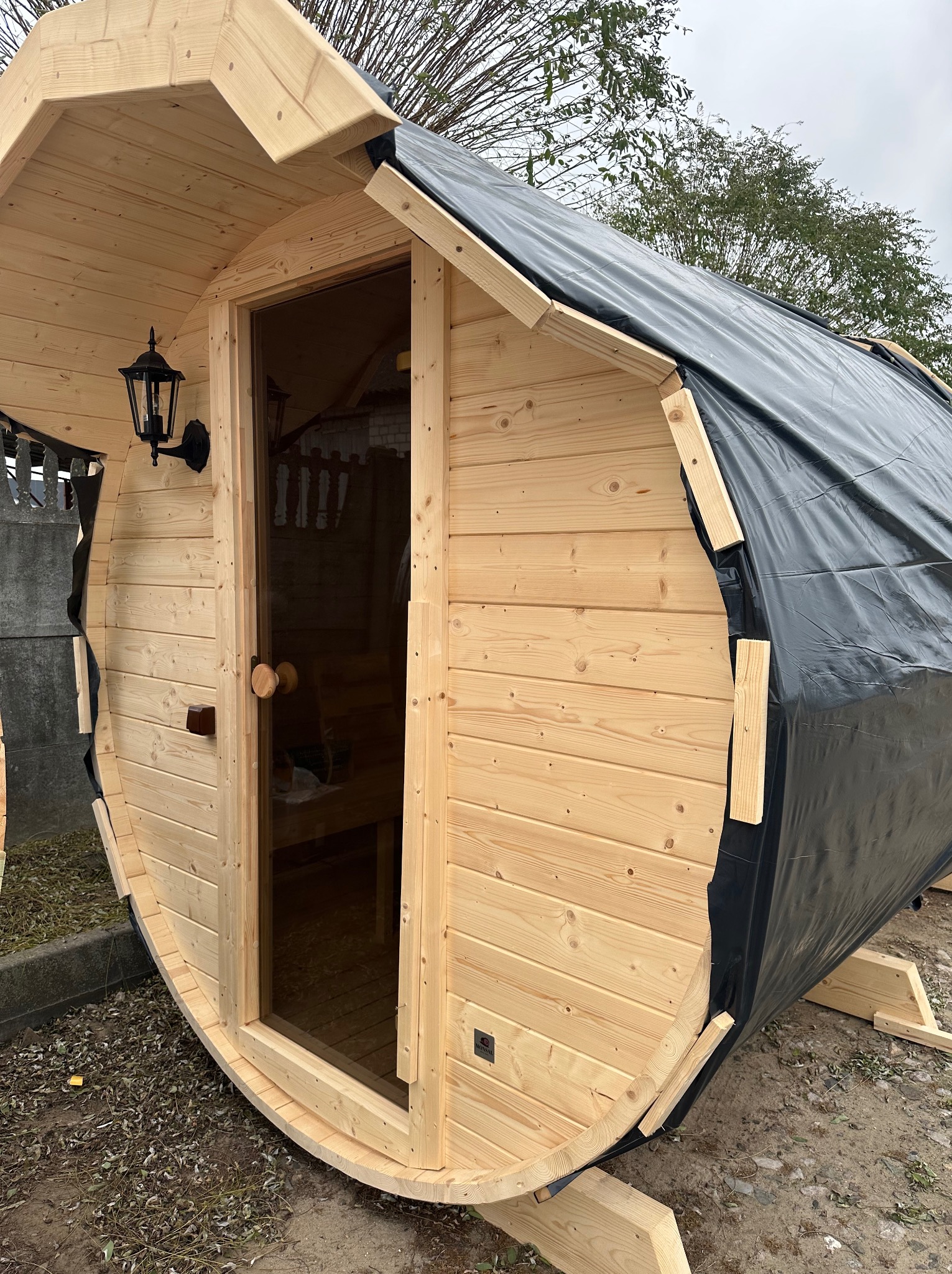 Drewniana sauna w kształcie beczki z czarną plandeką na części dachu, widoczna szklana szyba w drzwiach i lampa na ścianie zewnętrznej.