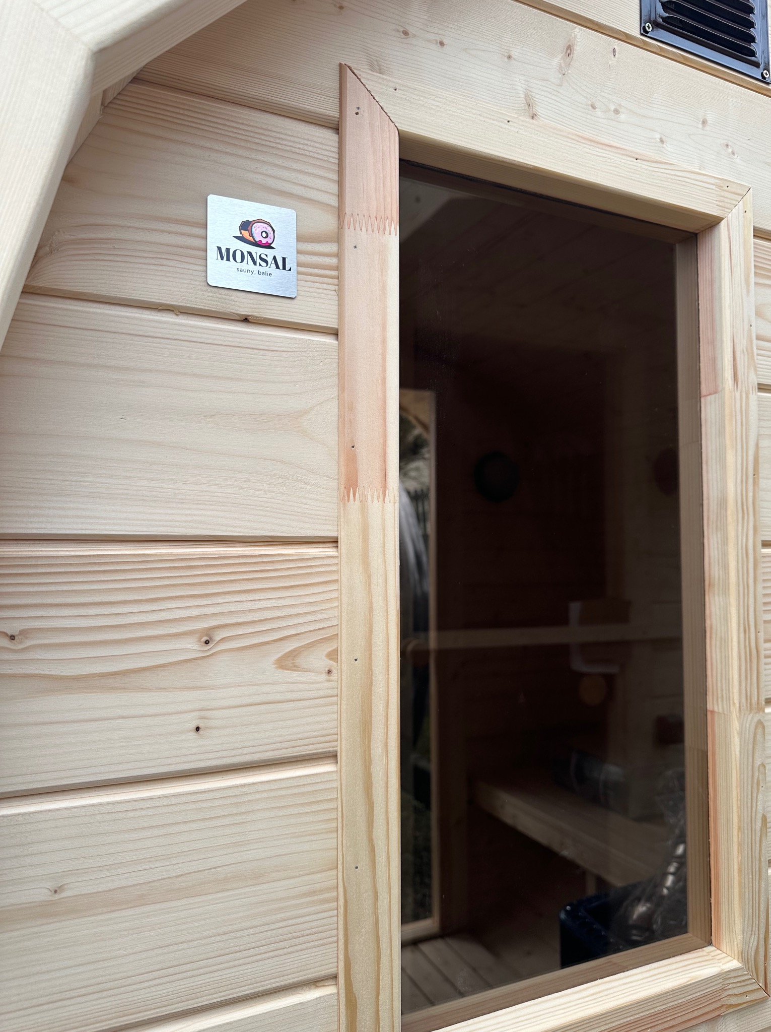 Fragment zewnętrznej ściany drewnianej konstrukcji z oknem, widoczne łączenia desek na wpust i wypust, oraz logo producenta przy górnej krawędzi