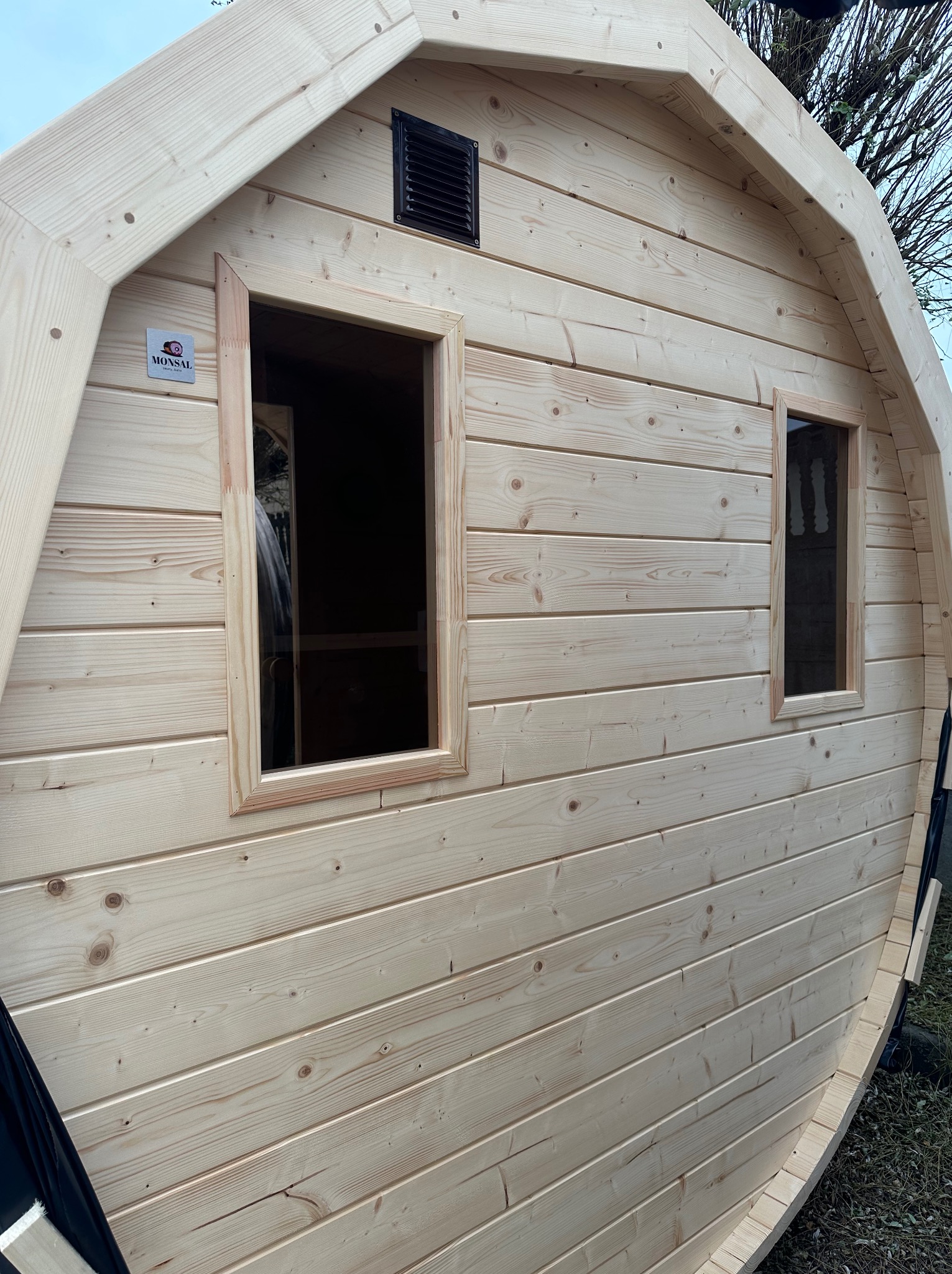 Zewnętrzny widok drewnianej sauny beczki z dwoma oknami i kratką wentylacyjną, widoczny znak producenta Monsal.