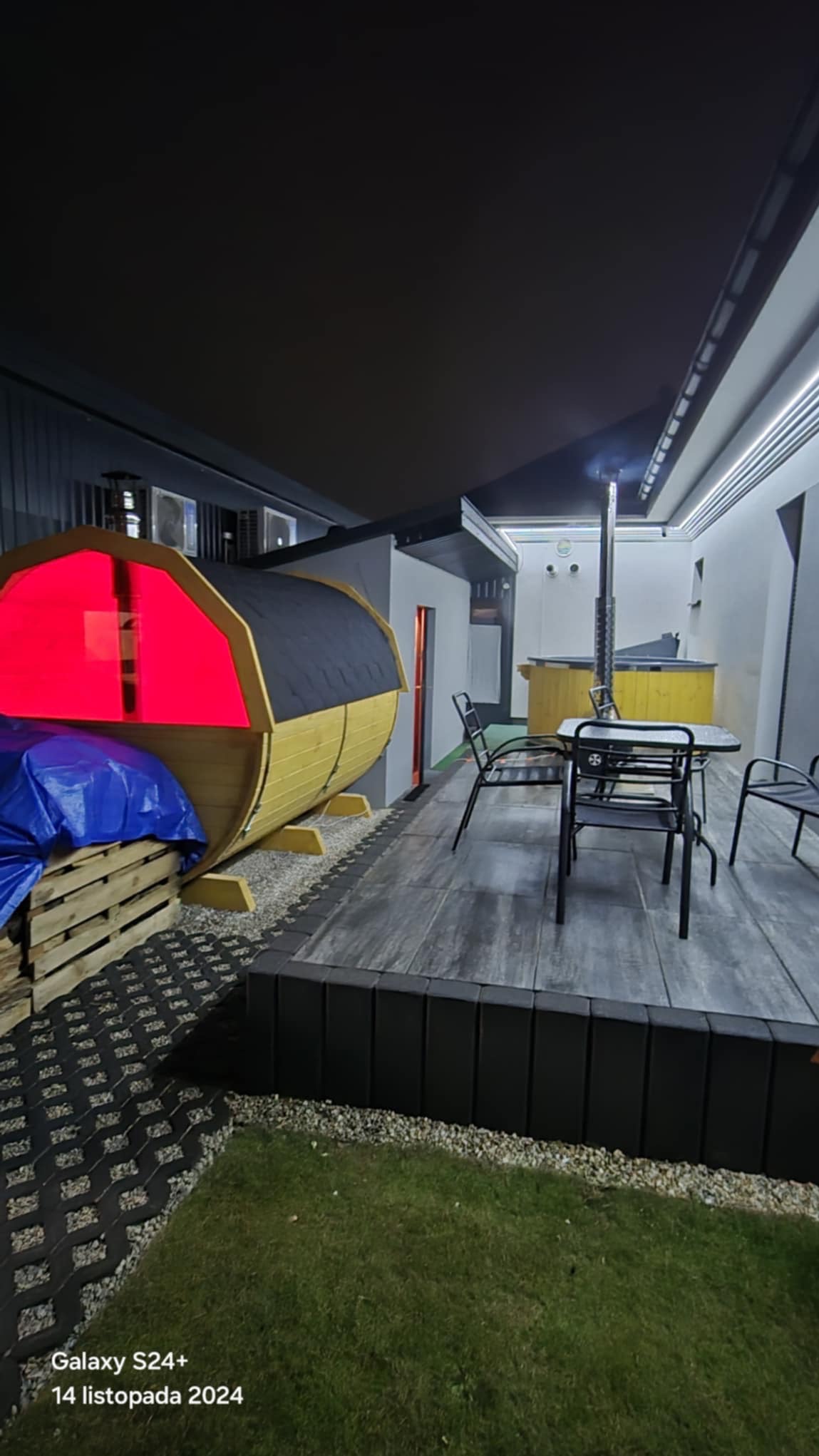 Drewniana sauna w kształcie beczki z czerwonym oświetleniem wewnętrznym, częściowo przykryta niebieską plandeką, obok zestaw mebli ogrodowych na tarasie i trawnik.