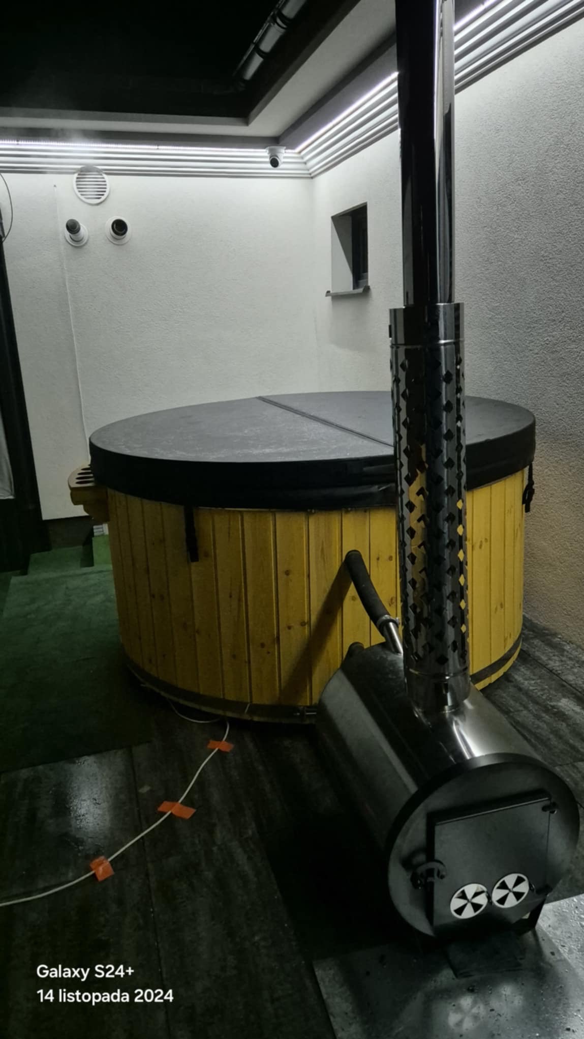 Drewniana balia z pokrywą i piecem opalanym drewnem, zainstalowana w pomieszczeniu w Łazach. Widoczny komin pieca i kabel na podłodze.