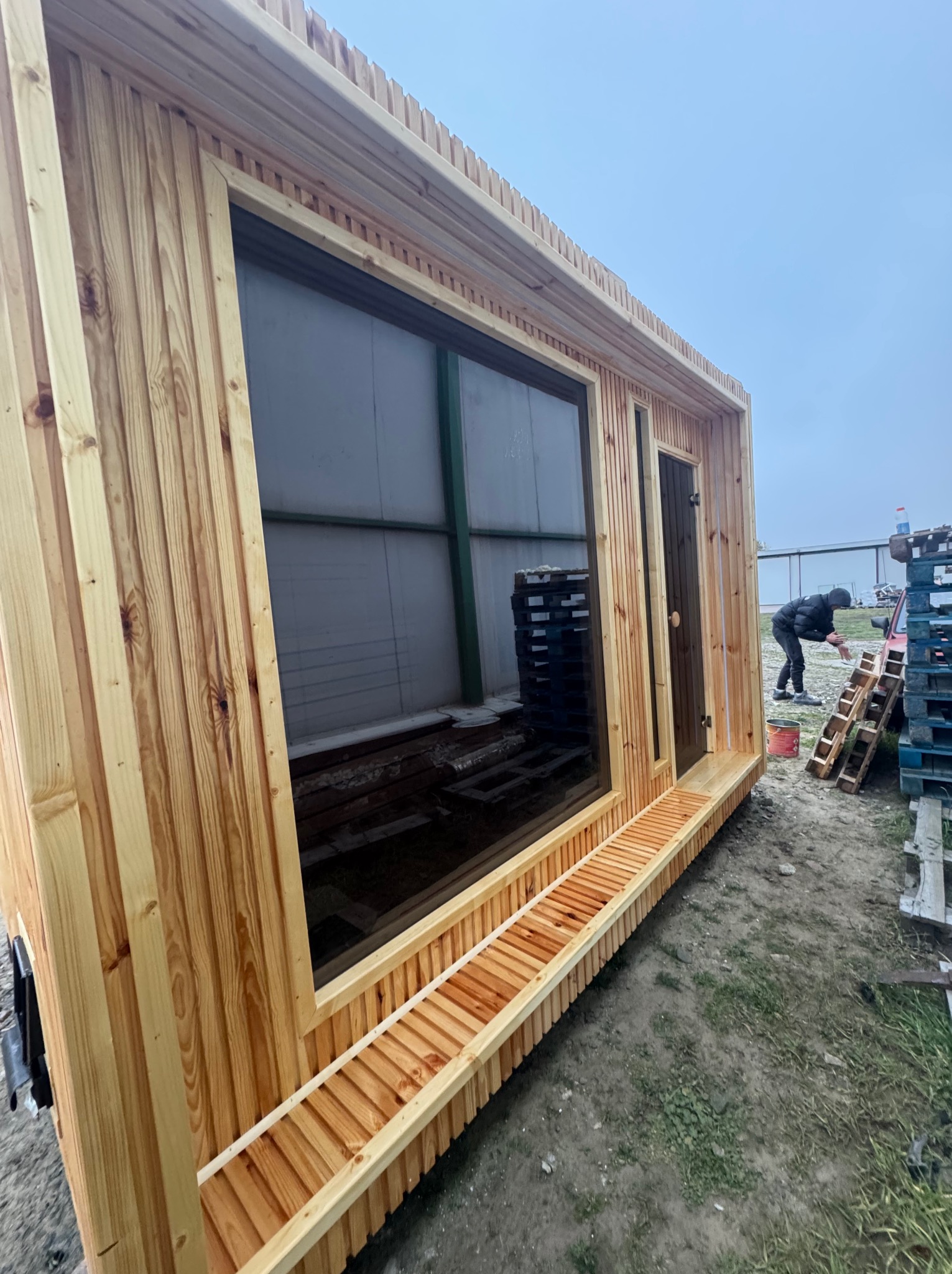 Drewniana konstrukcja z dużym oknem i małym wejściem, prawdopodobnie w trakcie budowy, widoczny pracownik malujący elementy obok, na zewnątrz, pochmurna pogoda.