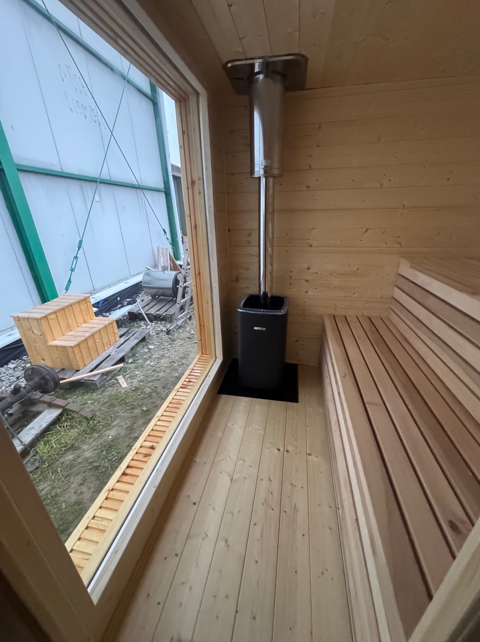 Wnętrze drewnianej sauny w trakcie budowy, widoczny piec Harvia z kominem, drewniane ławy i surowe otoczenie budowy za oknem.