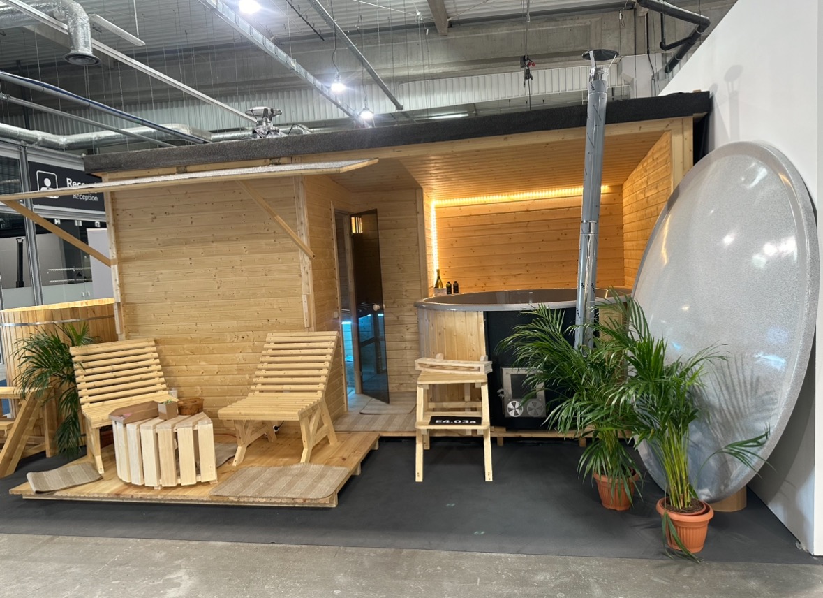 Drewniana sauna zewnętrzna z wanną, oświetleniem LED i kominem, z drewnianymi leżakami i stolikiem na podeście, prezentowana na ekspozycji targowej.