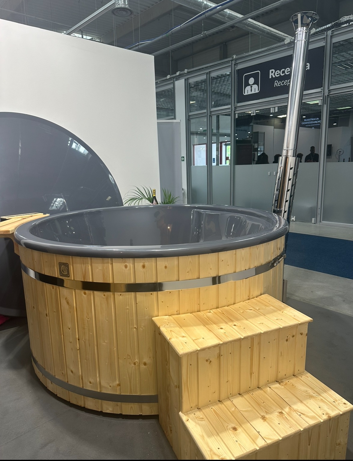 Drewniana balia z szarym wnętrzem i metalowym kominem, ustawiona na dwustopniowych schodkach z jasnego drewna, prezentowana w hali targowej z widocznym napisem 'Recepcja'.