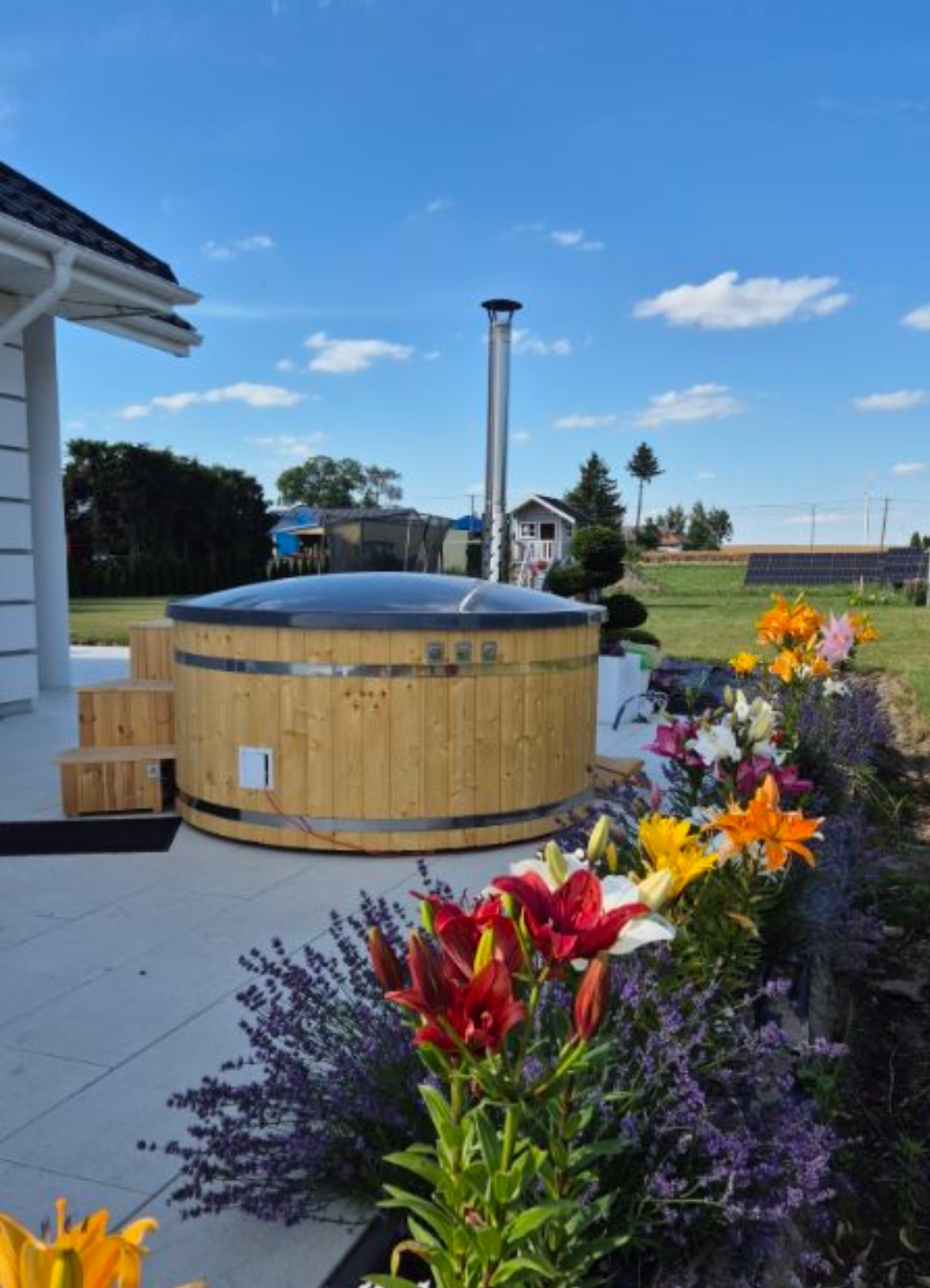 Drewniana balia z metalowymi obręczami i kominem, ustawiona na betonowym tarasie, z widocznymi schodkami i rabatą kwiatową z liliami i lawendą w tle, na tle błękitnego nieba.