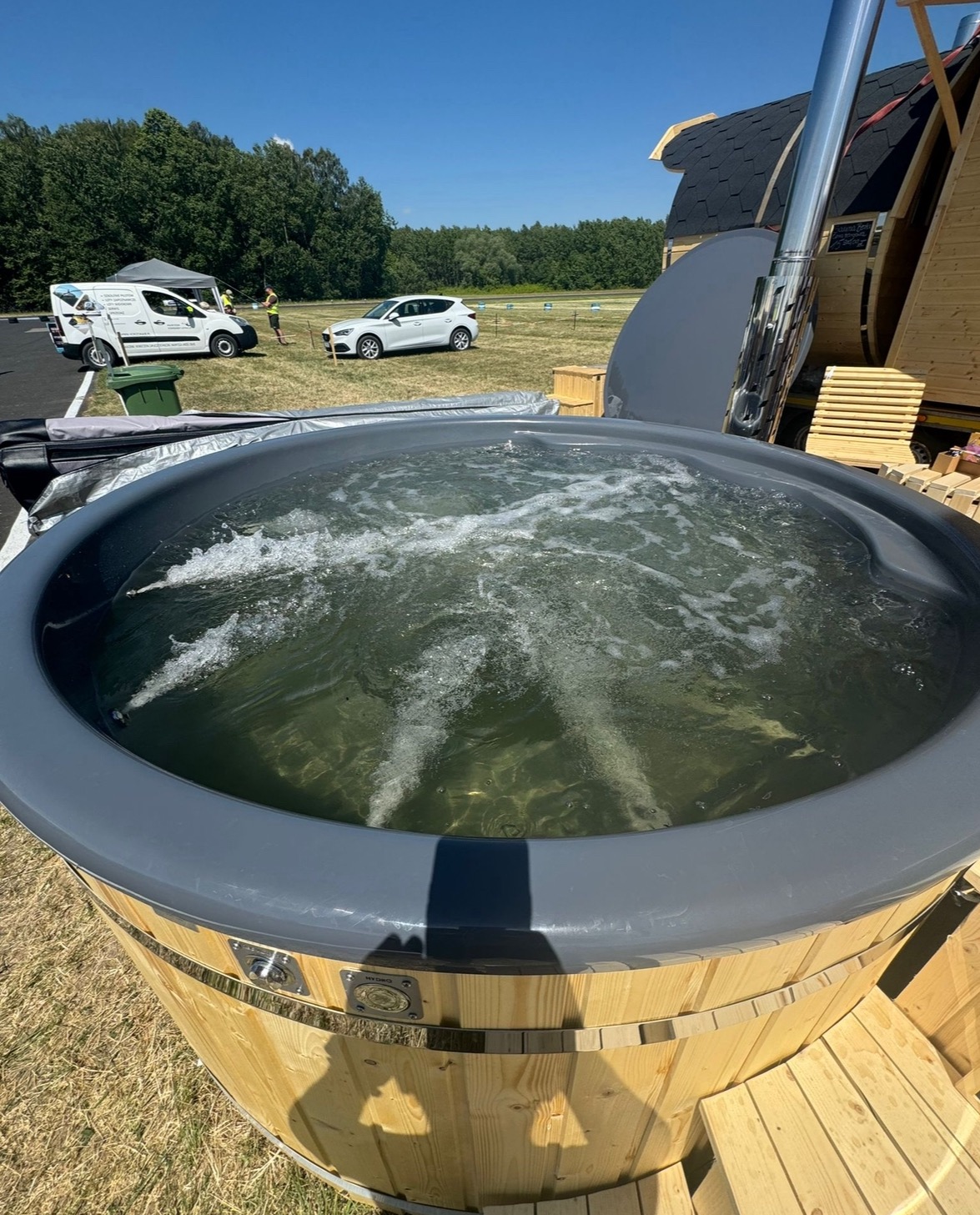 Drewniana balia z szarą obudową wypełniona wodą z bąbelkami, na zewnątrz w słoneczny dzień, w tle samochody dostawcze i osobowe oraz fragment sauny beczki z kominem.