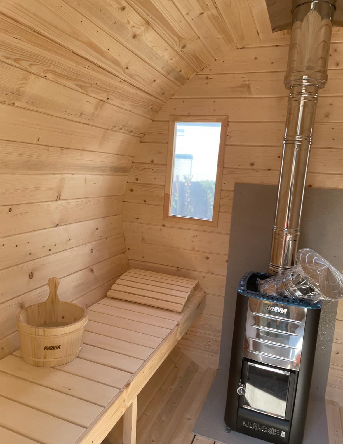 Wnętrze drewnianej, beczkowej kabiny z piecem Harvia, drewnianym wiadrem i małym oknem.