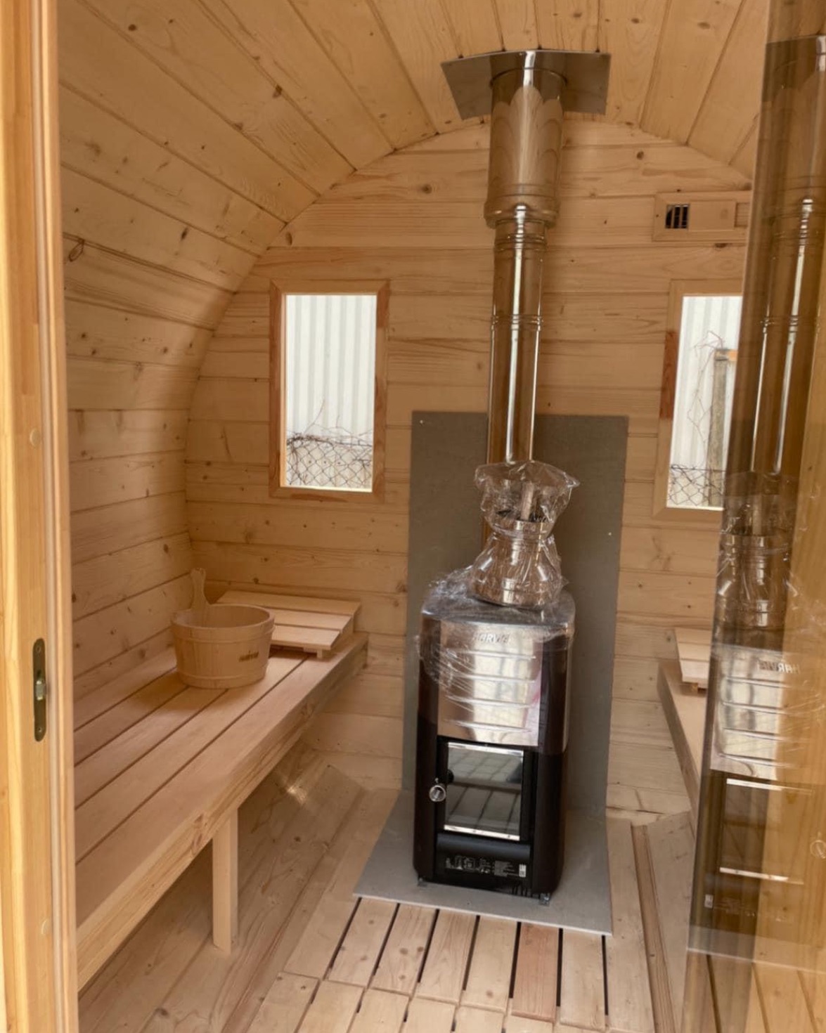 Wnętrze nowej, drewnianej sauny beczki z piecem Harvia jeszcze w folii ochronnej, widoczne drewniane ławki i wiadro.