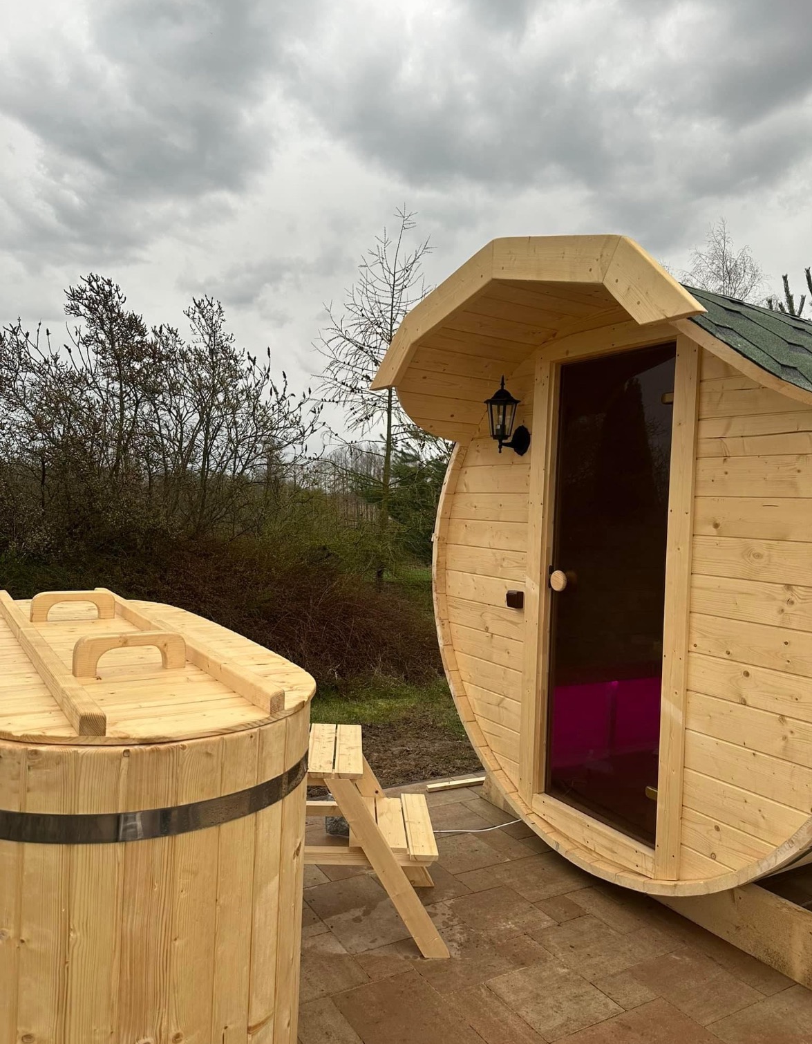 Drewniana beczka z pokrywą i zewnętrzna sauna beczka z małym gankiem i lampą ścienną, na tle pochmurnego nieba i drzew
