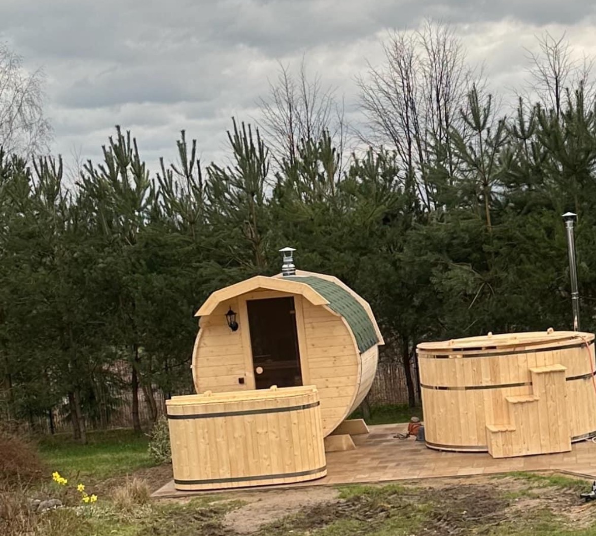Komplet drewnianych saun beczkowych i balii ogrodowych na wybrukowanym tarasie, z zielonym dachem sauny i tłem z wysokich sosen.