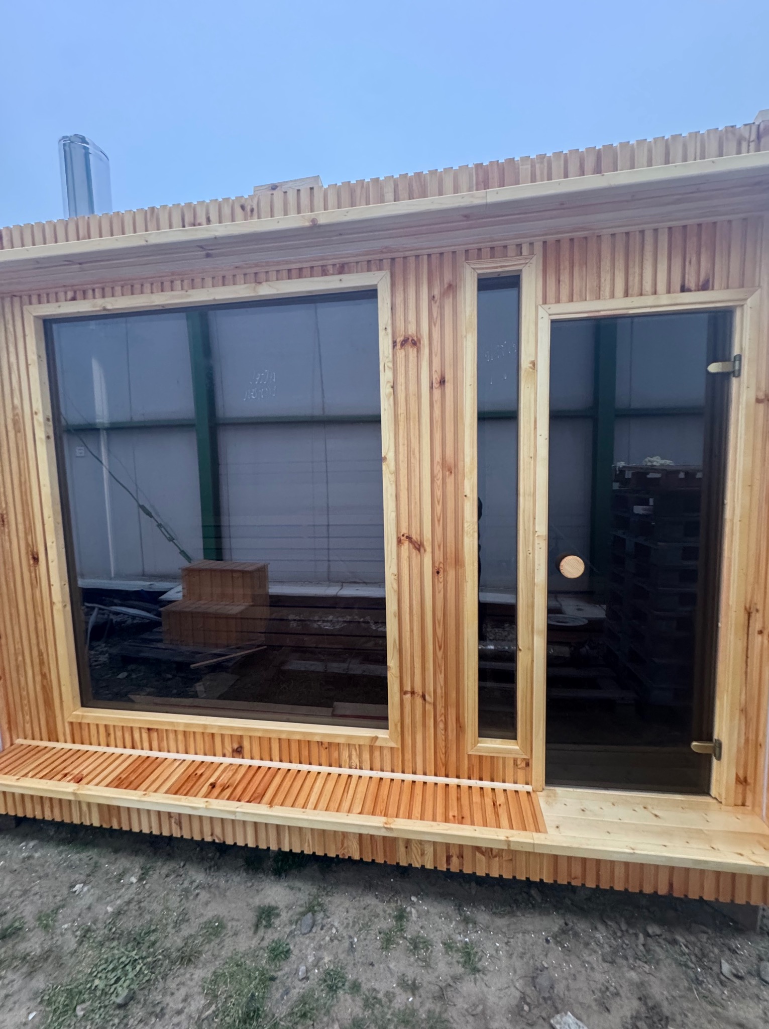 Surowa drewniana konstrukcja z dużym oknem i przeszklonymi drzwiami, prawdopodobnie w trakcie budowy, widoczne rusztowania i materiały budowlane w tle za oknem.