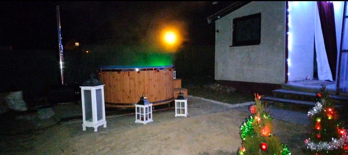 Oświetlona drewniana balia z podświetloną wodą na zewnątrz w nocy, otoczona lampionami i choinkami, w tle budynek.