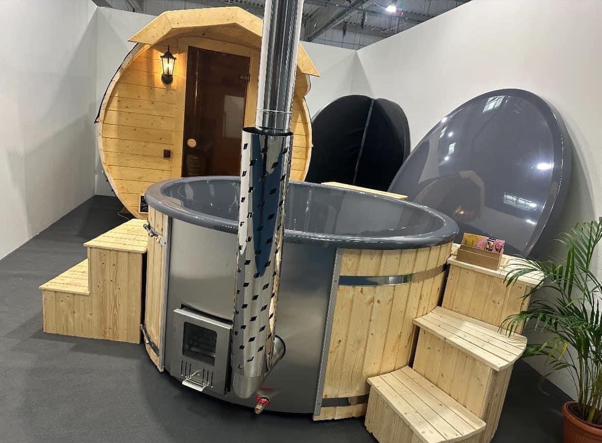 Wystawa: Beczka z szarą wkładką i piecem opalanym drewnem, schodki z surowego drewna, obok beczkowa kabina z oświetleniem zewnętrznym, w tle pokrywa.