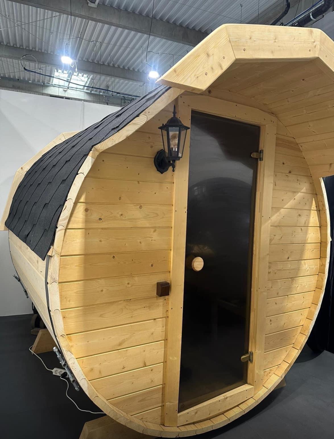 Drewniana, beczkowata konstrukcja z czarnym dachem z gontu bitumicznego i szklanymi drzwiami, oświetlona zewnętrzną lampą w stylu retro