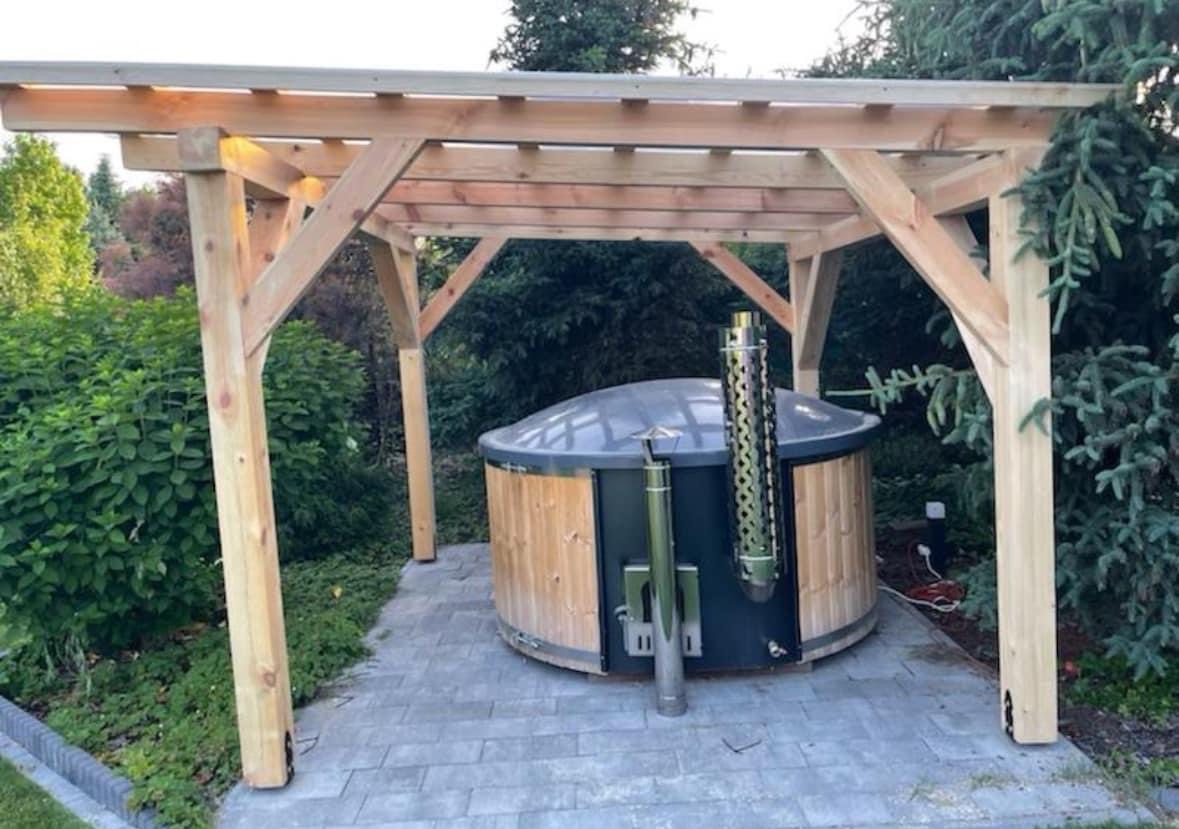 Drewniana sauna beczka z kominem pod prostą, jasną altaną z surowego drewna, na szarym brukowanym podłożu, otoczona zielenią ogrodu.