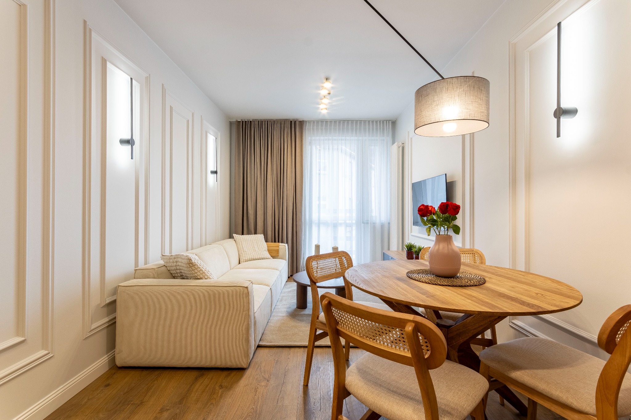 Jasne wnętrze mieszkania w stylu modern classic: sofa z kremowej tkaniny, okrągły drewniany stół z rattanowymi krzesłami, dekoracyjne panele ścienne i minimalistyczne oświetlenie.