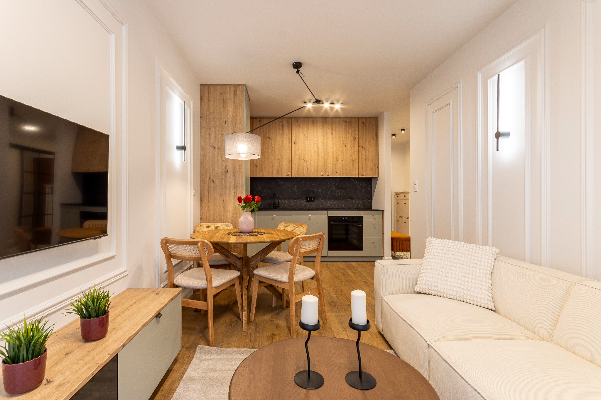 Jasne wnętrze mieszkania z drewnianym stołem jadalnym, rattanowymi krzesłami, sofą w odcieniach bieli i aneksem kuchennym z szarymi szafkami oraz czarnym blatem.