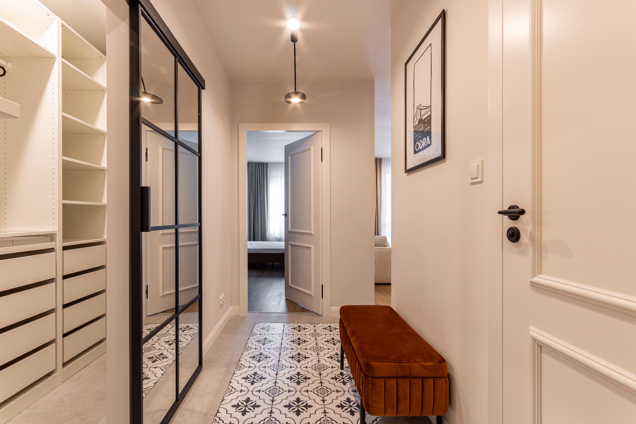 Korytarz z szafą wnękową za przesuwnymi drzwiami z czarnymi szprosami, wzorzysta podłoga, brązowa ławka i plakat na ścianie.