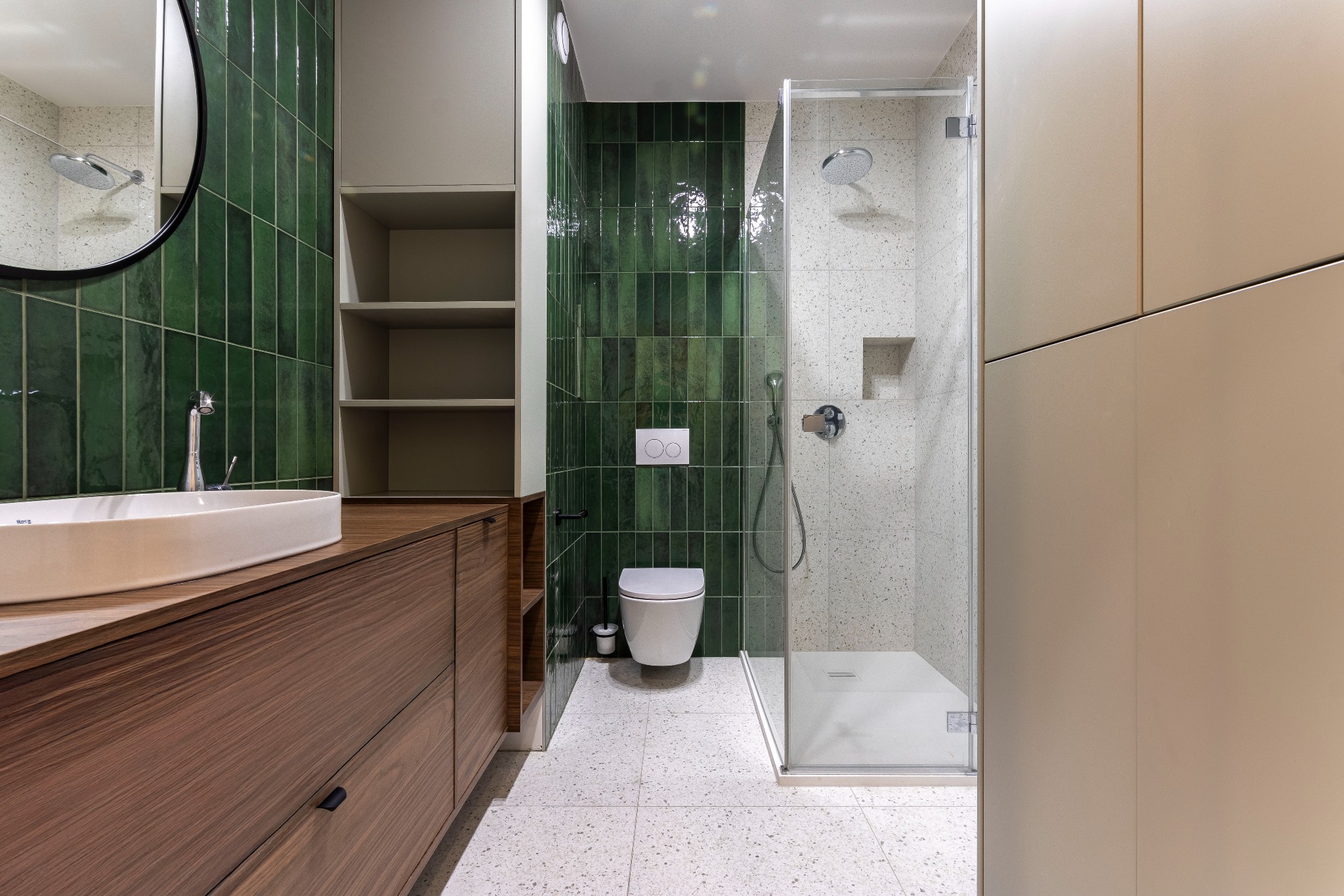 Nowoczesna łazienka z zielonymi, błyszczącymi płytkami ułożonymi pionowo, drewnianą szafką pod umywalką, białą umywalką nablatową, toaletą podwieszaną i kabiną prysznicową z deszczownicą.