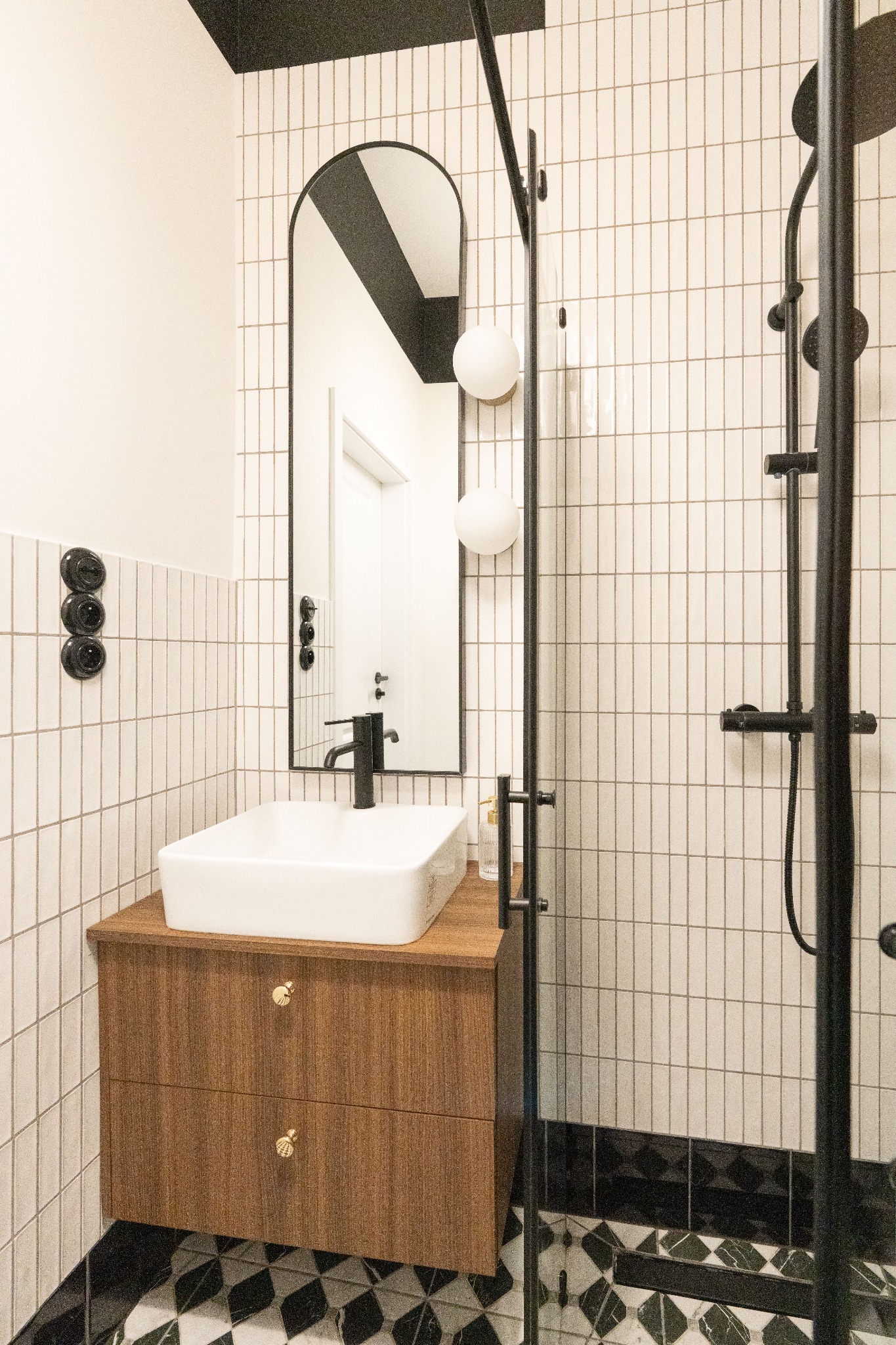 Nowoczesna łazienka: białe, pionowe płytki, drewniana szafka z umywalką, czarne akcenty (armatura, rama lustra), wzorzysta podłoga. Minimalistyczny design.