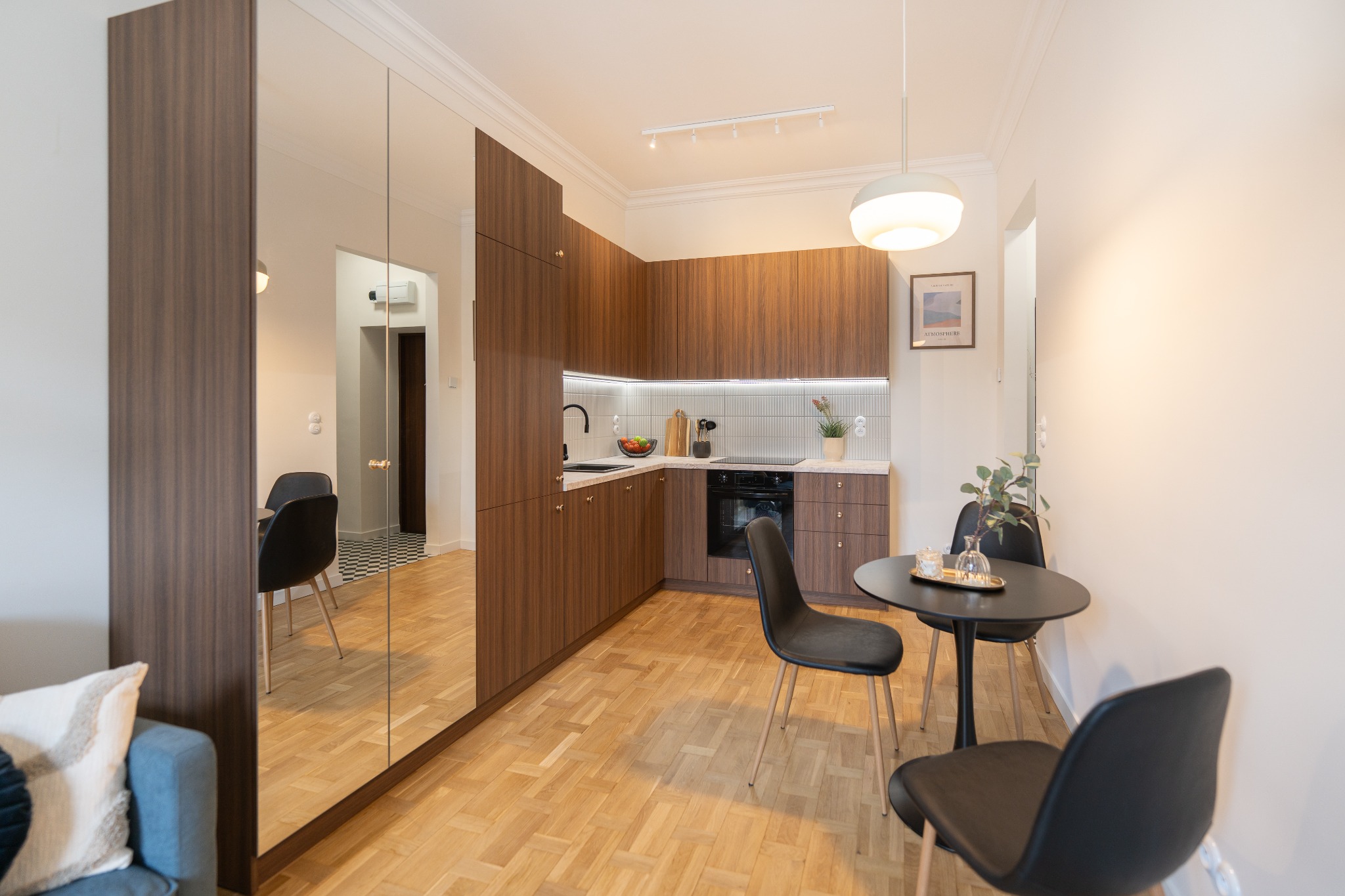 Nowoczesna kuchnia z drewnianymi szafkami i lustrem, odbijającym część jadalni z czarnym stołem i krzesłami. Parkiet w jodełkę. Oświetlenie punktowe i wisząca lampa.